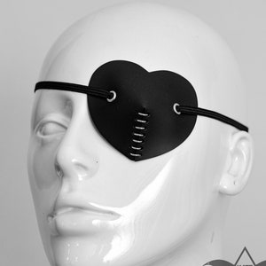 May include: A black leather heart-shaped eye patch with a stitched detail and two metal eyelets. The patch is worn on a white mannequin head.