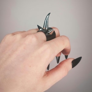 Könnte beinhalten: Ein schwarzer Ring mit silbernen Spikes. Der Ring wird an einem Finger mit schwarzem Nagellack getragen.