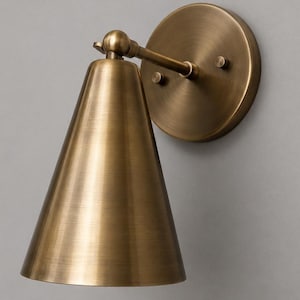 Op de afbeelding: Een messing wandlamp met een conische kap en een cirkelvormige achterplaat. De lamp heeft een warme, metallic afwerking en een eenvoudig, elegant ontwerp. De lamp is aan een grijze muur bevestigd.