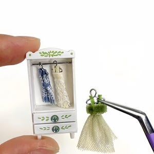 May include: Miniature white wardrobe with two drawers and green leaf accents. Inside, two tiny dresses hang on a rod. A third dress, held by tweezers, is light green with a mesh skirt. A charming dollhouse accessory.
