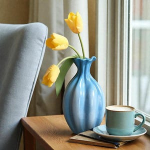 May include: A blue ceramic vase with vertical ridges holds three yellow tulips. A teal coffee cup and saucer sit on a wooden table next to the vase. The scene is set near a window with natural light.
