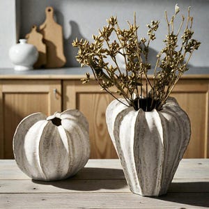 May include: Two white ceramic vases with a textured, ribbed design sit on a wooden table. One vase is filled with dried, golden-brown stems. The vases have a unique, organic shape, adding a touch of rustic elegance to the decor.