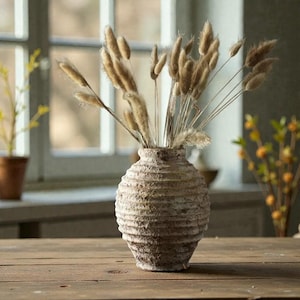 May include: A beige ceramic vase with a textured, ribbed design contains dried, tan ornamental grasses. The vase is on a wooden surface, with a window and other plants in the background. The vase is a decorative home accent.