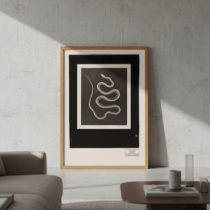 Op de afbeelding: Een ingelijste verticale wandkunstprint met een witte, kronkelende slangenillustratie gecentreerd op een zwarte rechthoekige achtergrond. De print zit in een lichthouten lijst en bevat een klein rechthoekig logo met tekst in de hoek.