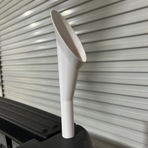 May include: A white, 3D-printed funnel with a wide, angled opening and a narrow base. The funnel is positioned on a black surface against a backdrop of a corrugated metal wall. The design is simple and functional.