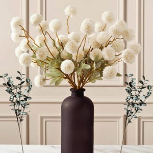 Peut inclure: Un vase marron foncé contient un bouquet de fleurs sphériques blanches avec des feuilles vertes et des tiges brunes. Deux brins de feuillage vert foncé flanquent le vase. L'arrangement est placé sur un fond neutre.