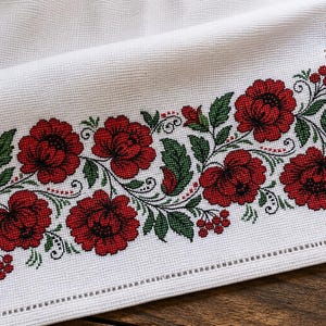Könnte beinhalten: Ein weißes rechteckiges Textil mit einer floralen Kreuzstichbordüre. Das Muster zeigt leuchtend rote Rosen mit dunkelgrünen Blättern und kleinen roten Beerenclustern, die als durchgehende horizontale Ranke auf dem Stoff angeordnet sind.