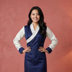 May include: A woman wearing a navy blue dress with white sleeves and a white collar. The dress has a floral pattern and a wrap-style neckline. The sleeves have navy blue cuffs. The woman is smiling and standing in front of a red wall.
