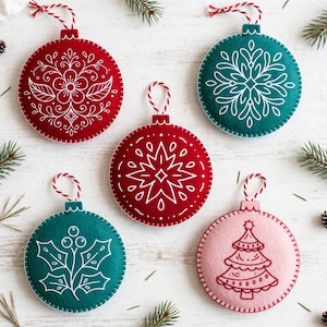 May include: Five circular felt ornaments with white embroidered designs. Two are red, two are teal, and one is light pink. The designs include a flower, a starburst, holly leaves with berries, and a Christmas tree. Each has a red and white string.