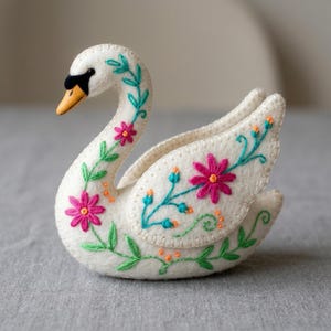 May include: A white felt swan figurine featuring hand-stitched embroidery. The swan has a black and orange beak and is decorated with vibrant pink flowers, green leafy vines, and small orange dots stitched along its neck and wings.