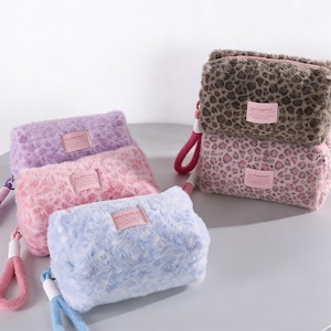 May include: A collection of fluffy, rectangular cosmetic bags in various colors: lavender, pink, light blue, and two with leopard print. Each bag has a matching wrist strap and a small rectangular label.