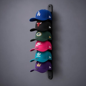 Könnte beinhalten: Ein vertikaler, schwarzer Wandhalter mit sechs übereinander angeordneten Baseballkappen. Die Kappen sind blau mit weißem LA-Logo, schwarz mit rotem Stier-Logo, dunkelgrün mit G-Logo, pink mit weißem Nike-Logo, türkis mit weißem Adidas-Logo und lila mit goldenem Kronen-Logo.