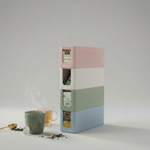 Puede incluir: Una pila vertical de cuatro cajas de almacenamiento rectangulares de plástico con un patrón de panal texturizado en rosa pastel, blanco, verde salvia y azul claro. Cada caja contiene una caja de té. Una taza de cerámica con té humeante y una cuchara dorada están cerca.