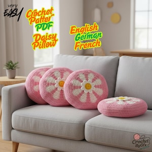 Puede incluir: Cuatro cojines de crochet rosa con forma de margarita sobre un sofá gris. Los cojines tienen pétalos blancos y centros amarillos. El texto de la imagen dice "very EASY Crochet Patter PDF Daisy Pillow" y "English German French".