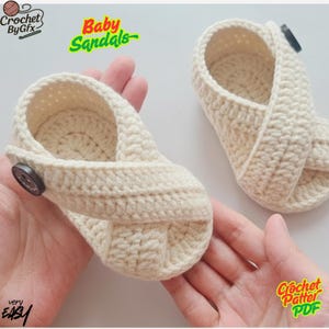 May include: A pair of cream-colored, crocheted baby sandals. The sandals feature a criss-cross strap design and a button closure. The text "Baby Sandals" is in green and yellow. The text "Crochet By Gfx" and "very EASY" are also visible.
