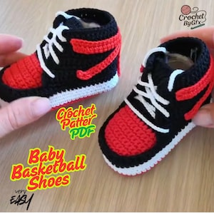 Può includere: Un paio di scarpine da neonato all'uncinetto, ispirate alle scarpe da basket. Le scarpe sono nere e rosse, con lacci e suole bianche. Il testo "Baby Basketball Shoes" e "Crochet Pattern PDF" è visibile.