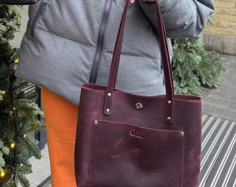 May include: Two leather tote bags are displayed. One is black, the other is a deep burgundy. Both have a rectangular shape, a front pocket, and long handles. The bags are held up in front of a person wearing a gray jacket and orange pants.