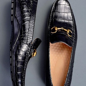 Pode incluir: Um par de mocassins pretos com cabedal de couro brilhante com relevo de crocodilo e um detalhe de bridão de metal dourado no peito do pé. O interior do sapato é forrado com um material bege claro e a sola tem um rasto de borracha preto.