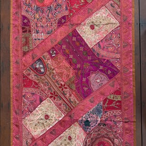 May include: A rectangular wall hanging featuring a patchwork of pink, magenta, and cream fabric panels. The textile is decorated with intricate embroidery, metallic thread stitching, beadwork, and sequins arranged in various geometric shapes.