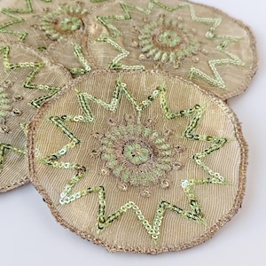May include: A set of circular coasters made of sheer beige fabric with metallic gold edges. Each coaster features a central embroidered floral motif in light green thread, surrounded by a zigzag pattern of light green sequins.