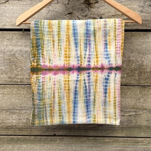 May include: A rectangular scarf featuring a tie-dye pattern with vertical stripes in shades of mustard yellow, blue, and pink on a cream background, with a horizontal dark green band across the centre, all displayed on a wooden hanger.