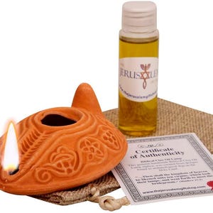May include: An orange clay oil lamp with a lit flame, a bottle of oil, and a certificate of authenticity. The lamp has an ornate design and sits on a burlap sack. The bottle has a white cap and a label with text.