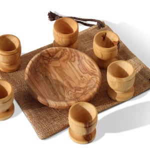 May include: A wooden breakfast set comprising a small bowl and six egg cups. The set is crafted from light-coloured wood with a natural grain pattern. The items are arranged on a burlap sack, with a drawstring closure visible.