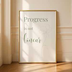 May include: A framed print with the words "Progress is not linear" in green script. The print has a gold frame and a minimalist floral illustration. The background is a soft, neutral color, and the print is displayed against a wall.