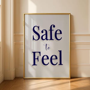 Pode incluir: Uma impressão de arte de parede retangular numa moldura dourada fina com o texto azul escuro Safe to Feel centrado num fundo cinzento claro. A tipografia utiliza um tipo de letra serifado a negrito para as palavras Safe e Feel com um tipo de letra script mais pequeno para to.