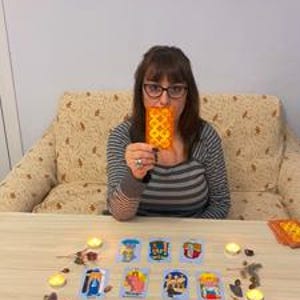May include: A person holding a tarot card in front of their face, with a table in front of them. The table is covered with tarot cards, small candles, and decorative stones. The person is wearing glasses and a striped shirt.