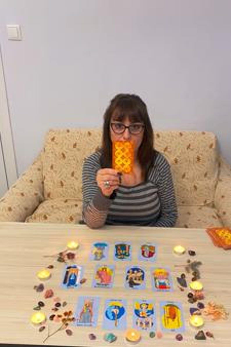 May include: A person holding a tarot card in front of their face, with a table in front of them. The table is covered with tarot cards, small candles, and decorative stones. The person is wearing glasses and a striped shirt.