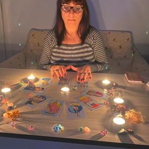 May include: A woman wearing glasses and a striped shirt sits at a table with tarot cards, lit candles, and crystals. The scene is dimly lit, creating a mystical atmosphere. The cards are spread out, suggesting a reading.