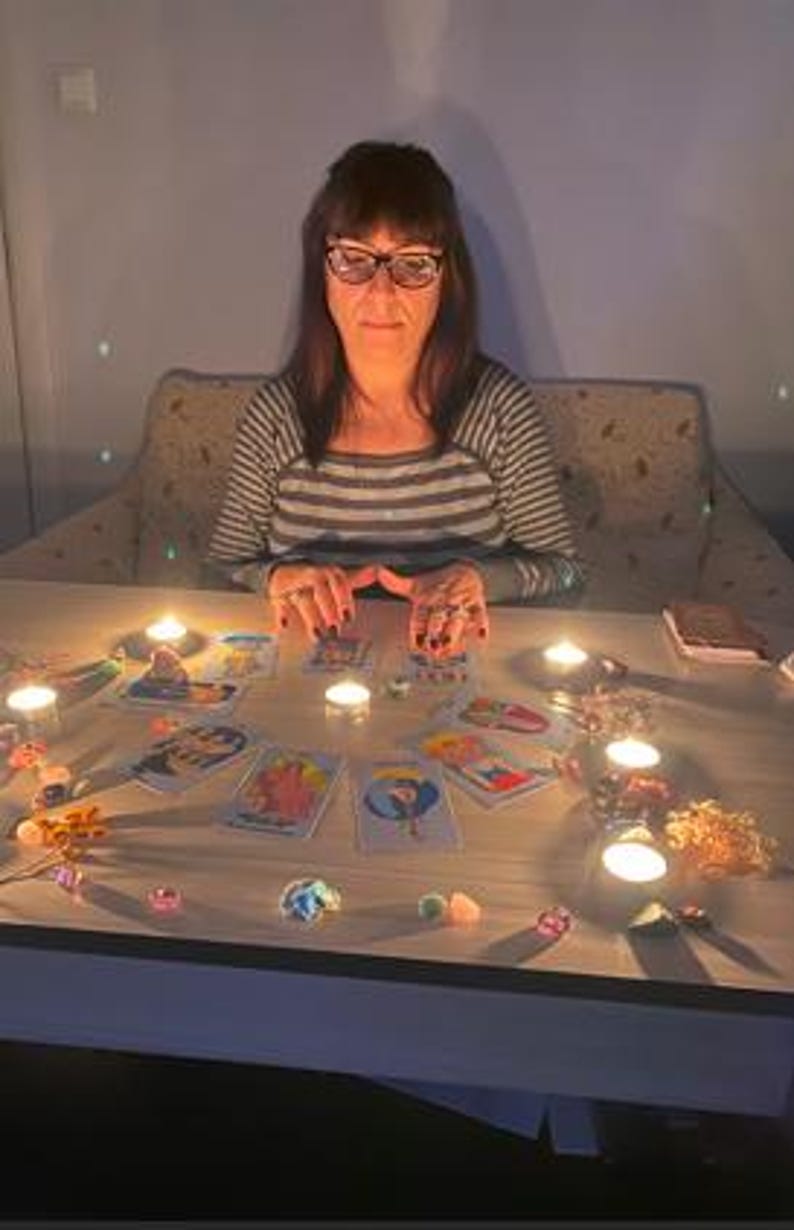 May include: A woman wearing glasses and a striped shirt sits at a table with tarot cards, lit candles, and crystals. The scene is dimly lit, creating a mystical atmosphere. The cards are spread out, suggesting a reading.