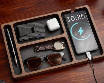 May include: A wooden desk organizer with compartments holding a pen, wallet, wireless earbuds, watch, sunglasses, and a smartphone charging at 65%. The phone displays the time as 10:25. The organizer is on a dark wood surface.