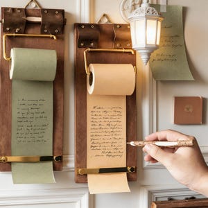 Puede incluir: Dos pizarras de desplazamiento de madera con herrajes de latón. Una pizarra tiene un rollo de papel verde, la otra un rollo de papel marrón, ambos con texto manuscrito. Una mano sostiene un bolígrafo cerca del papel marrón.