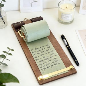 May include: A wooden clipboard with a roll of green paper and gold accents. The paper has handwritten text. A black pen and a lit candle are also visible on the white surface.