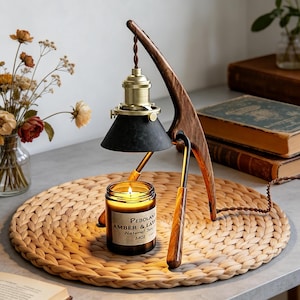 Könnte beinhalten: Eine Holz-Schreibtischlampe mit schwarzem Schirm und Messingakzenten. Darunter steht eine brennende Kerze in einem Glas. Das Etikett der Kerze lautet "PEBOLAN AMBER & LAVENDER NATURAL SOY 3.4OZ". Die Lampe steht auf einer gewebten Matte.