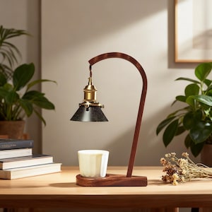 May include: A wooden desk lamp with a curved, dark brown frame. The lamp features a black lampshade with a brass fitting and a small white cup. The lamp is on a wooden surface with plants in the background.