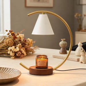 Puede incluir: Un calentador de velas dorado en forma de arco con una pantalla blanca. Una vela encendida en un frasco de vidrio se encuentra sobre una base de madera. El calentador está sobre una mesa con flores secas y objetos decorativos.