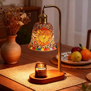 Puede incluir: Lámpara de mesa decorativa con pantalla de cristal de mosaico multicolor. La lámpara tiene una estructura metálica de color latón y una base de madera con una vela encendida. Un jarrón con flores secas y un plato de fruta también están sobre la mesa.