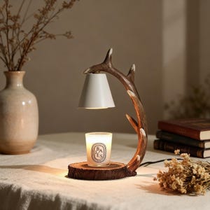 May include: A candle warmer featuring a white lampshade and a brown antler-shaped frame. The warmer is set on a wooden base, with a lit candle in a glass jar beneath the lampshade. A vase with dried flowers is in the background.