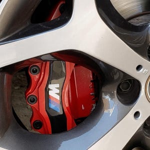 Peut inclure: Gros plan sur un étrier de frein de voiture rouge vif avec le logo BMW M en blanc, bleu et bleu clair. L'étrier est monté derrière une jante en alliage argent et gris. L'image met en évidence le design axé sur la performance.