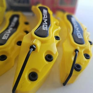 Puede incluir: Pinzas de freno AMG amarillas brillantes con detalles en negro y el logotipo AMG. Las pinzas tienen un diseño moderno con cabezas de pernos visibles y un tubo negro curvado. Piezas de automóviles.