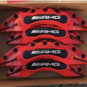 Puede incluir: Cuatro pinzas de freno AMG rojas en una caja de cartón. Cada pinza tiene el logotipo AMG en blanco sobre un fondo negro. Las pinzas son de color rojo brillante y parecen nuevas.