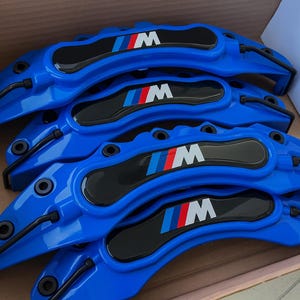May include: Four bright blue car brake calipers with black accents and the "M" logo in white, red, and blue. The calipers are arranged in a cardboard box, suggesting they are new and ready for installation.