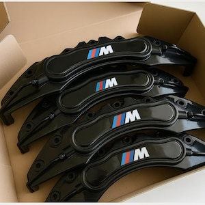 Pode incluir: Um conjunto de pinças de freio automotivas pretas com o logotipo BMW M no centro. O logotipo apresenta a letra "M" em branco, com listras vermelhas, azuis e azul claro. As pinças estão dispostas dentro de uma caixa de papelão.