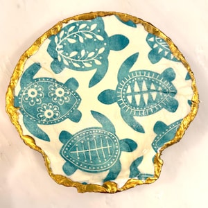 May include: A decorative seashell-shaped dish with a gold-painted edge. The interior features a cream-colored fabric printed with teal sea turtles in various designs. The dish is likely used for decorative purposes or as a small trinket tray.