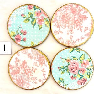 May include: Set of four round coasters with floral designs. Each coaster features a gold-painted edge. Two coasters have a light blue background with pink roses and green leaves. The other two have a white background with pink floral patterns.