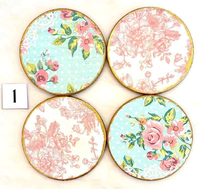May include: Set of four round coasters with floral designs. Each coaster features a gold-painted edge. Two coasters have a light blue background with pink roses and green leaves. The other two have a white background with pink floral patterns.
