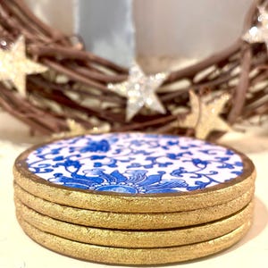 May include: A stack of four round coasters. Each coaster features a white background with a blue floral design and a gold-colored edge. The coasters are stacked on top of each other.